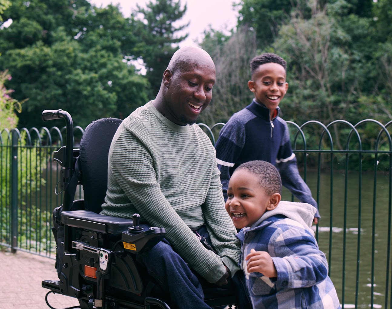 A man in a wheelchair plays with his two sons beside a lake in a park