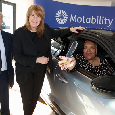 A smiling man and woman stand next to another woman sitting in a shiny new car holding a big key